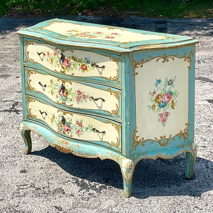 an old dresser painted with flowers and birds on it's sides is sitting in the gravel