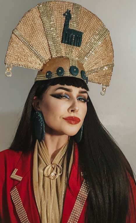 a woman with long black hair wearing a red jacket and gold headdress while standing next to a white wall