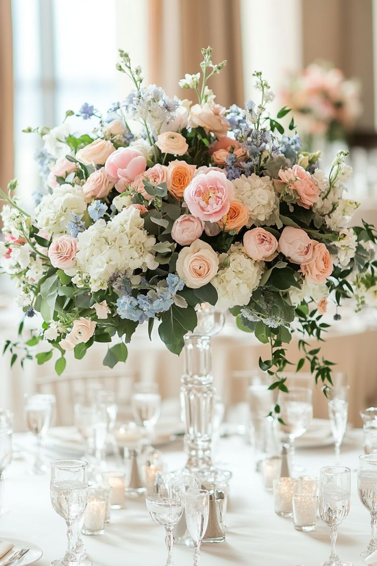 a tall vase filled with lots of flowers on top of a white table covered in wine glasses