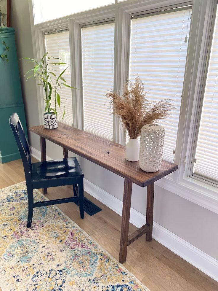 a wooden table sitting next to a window with blinds on the windowsill and a chair in front of it