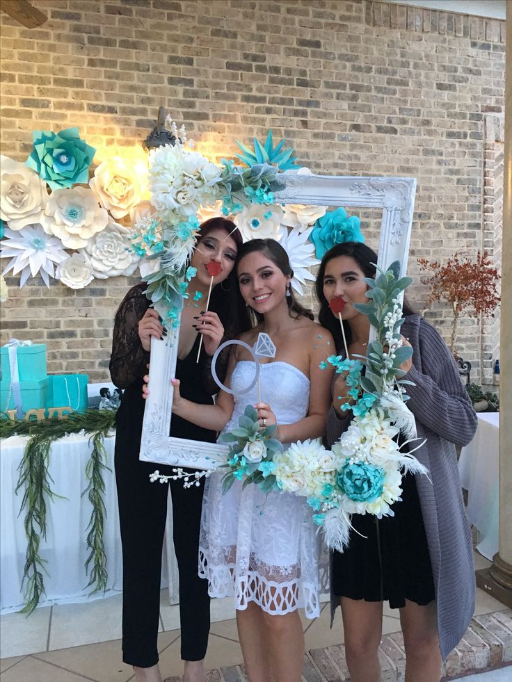 three women standing in front of a mirror with flowers and greenery around them, one holding a photo frame