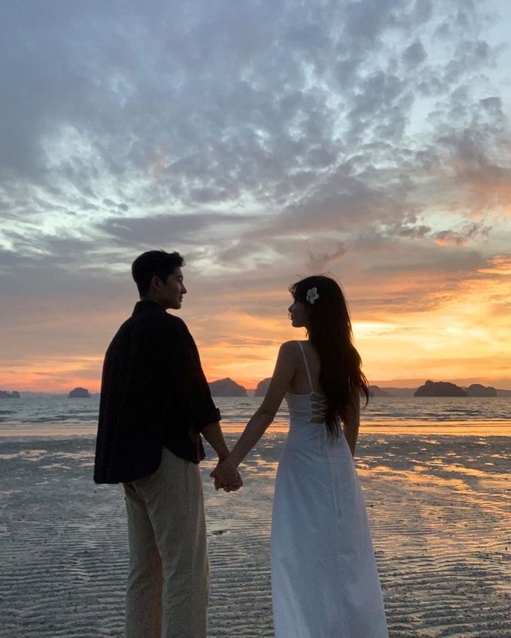 a man and woman holding hands while standing on the beach at sunset, with clouds in the background