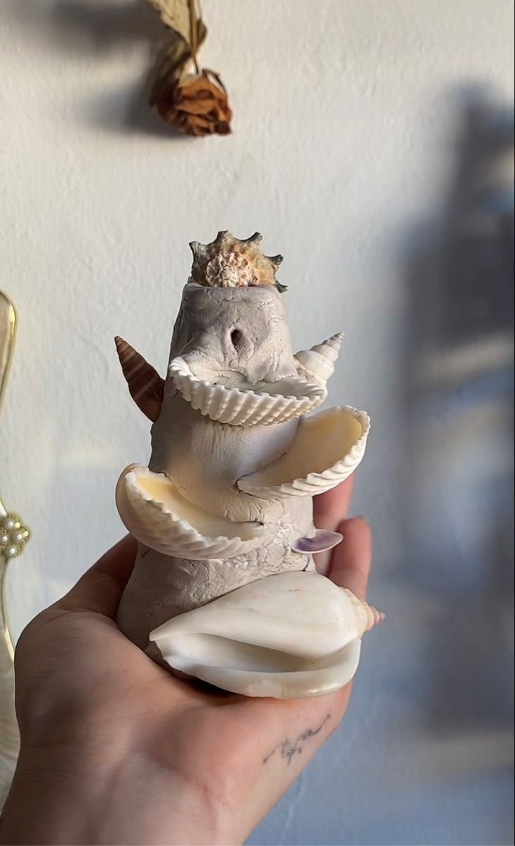 a hand holding an animal made out of shells and seashells with other items hanging on the wall in the background