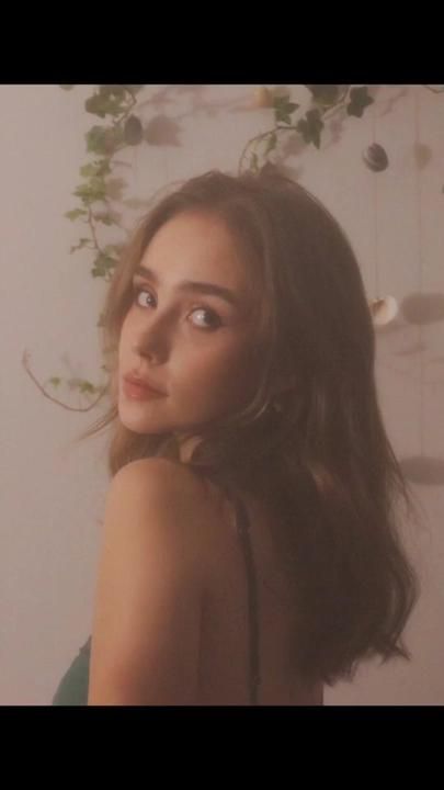 a woman with long brown hair standing in front of a white wall and greenery