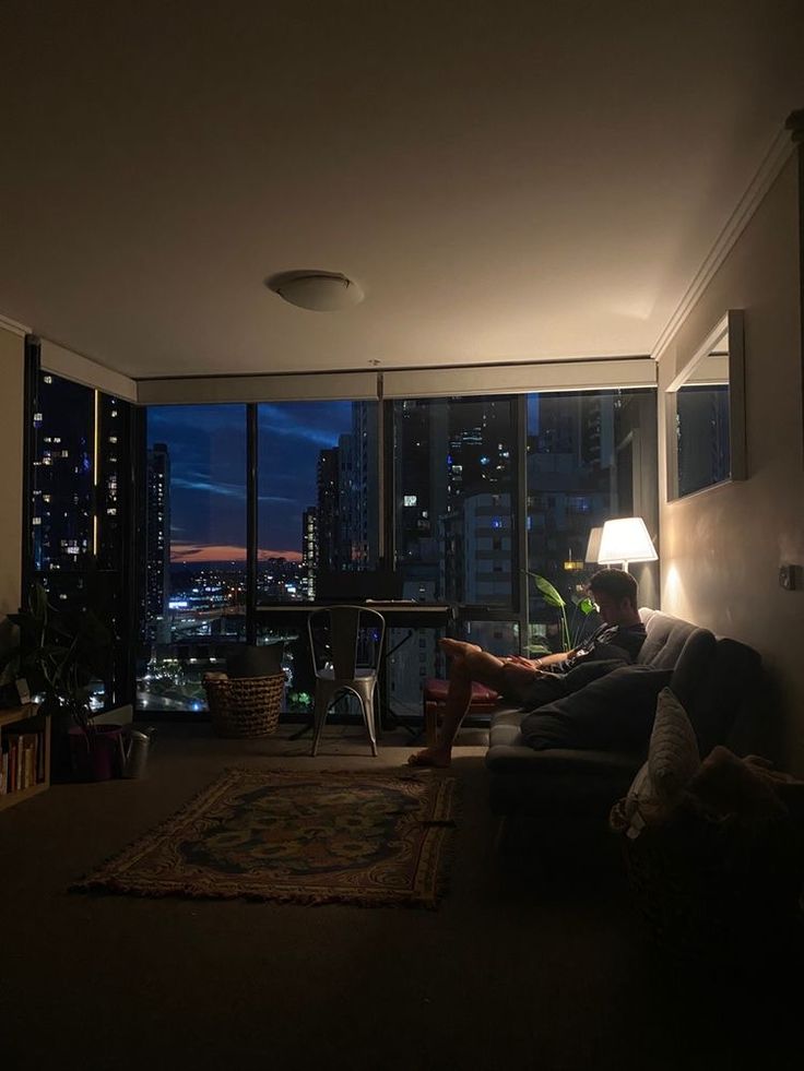 a man sitting on a couch in front of a window with the city lights lit up