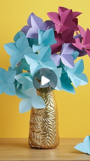 a vase filled with paper flowers sitting on top of a wooden table in front of a yellow wall