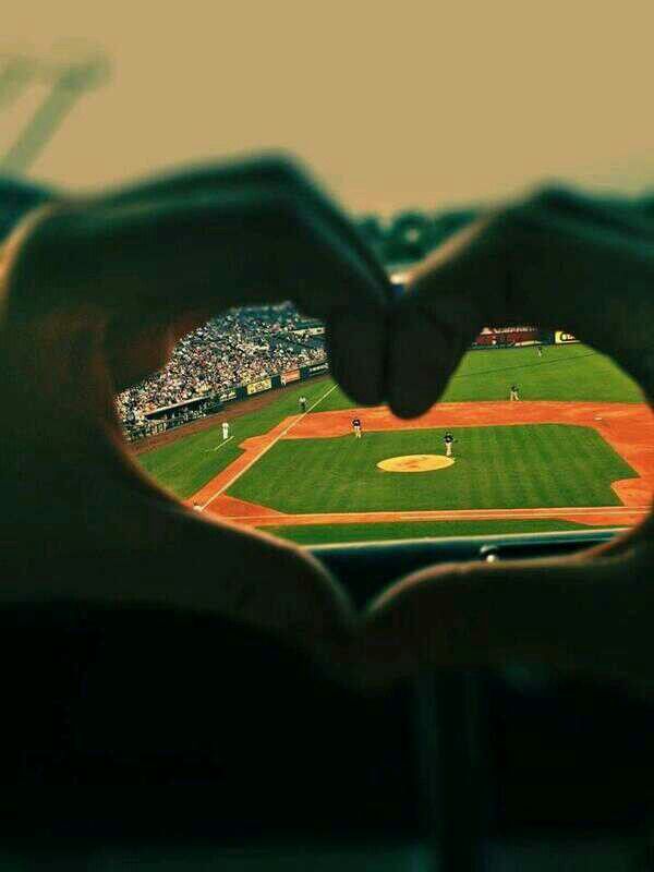 a baseball field seen through a heart shaped hole in the middle of two hands, with a stadium in the background