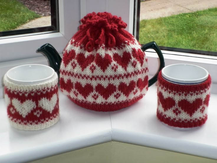 a red and white knitted tea pot next to two mugs