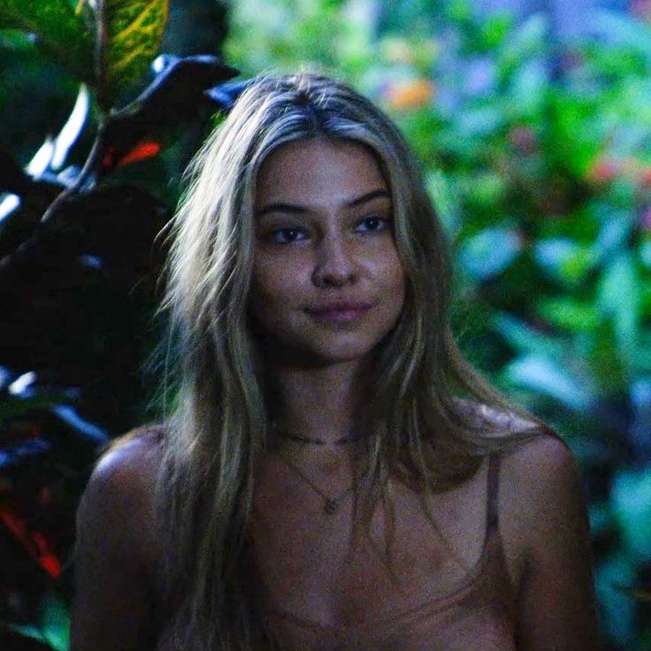 a woman with no shirt standing in front of some plants and trees, looking at the camera