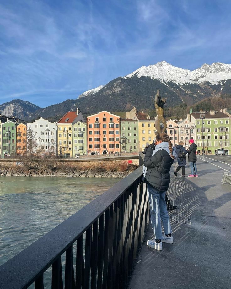 people standing on a bridge looking at the water and buildings in the background with snow - capped mountains behind them