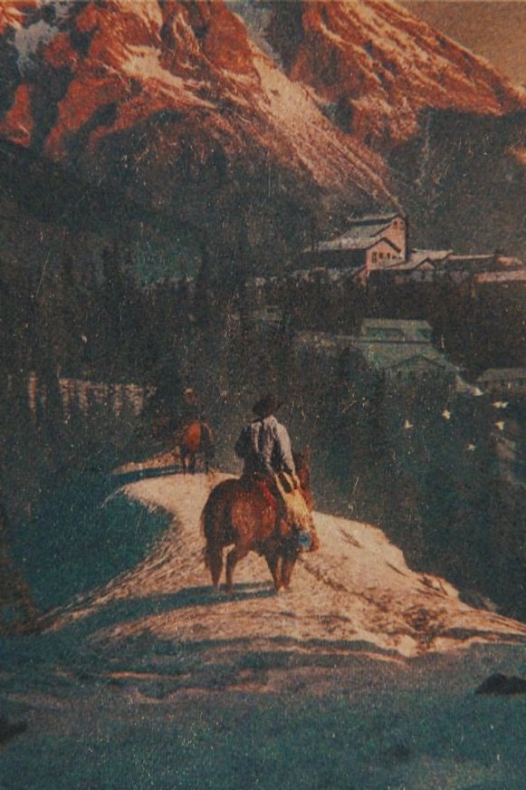 two horses walking down a road with mountains in the backgrouds and houses on either side