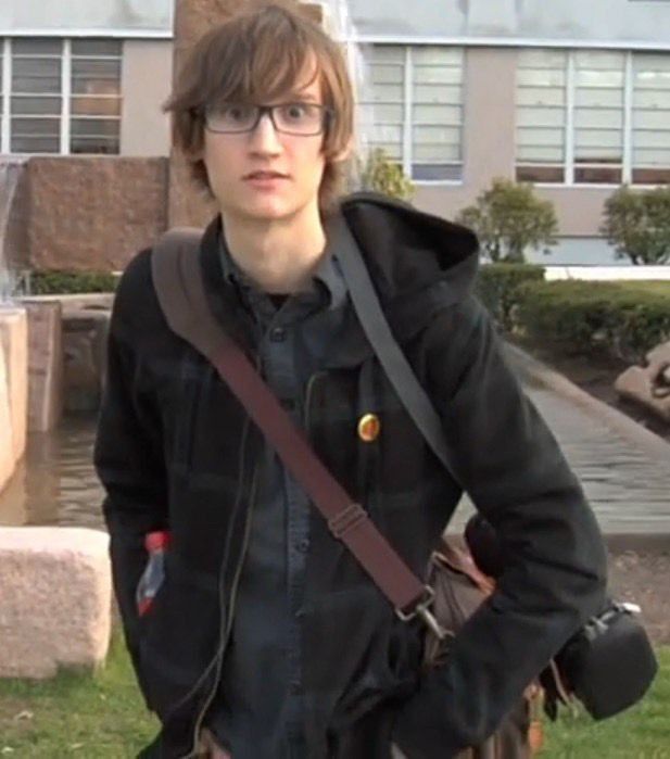 a young man with glasses and a backpack standing in front of a building, looking at the camera