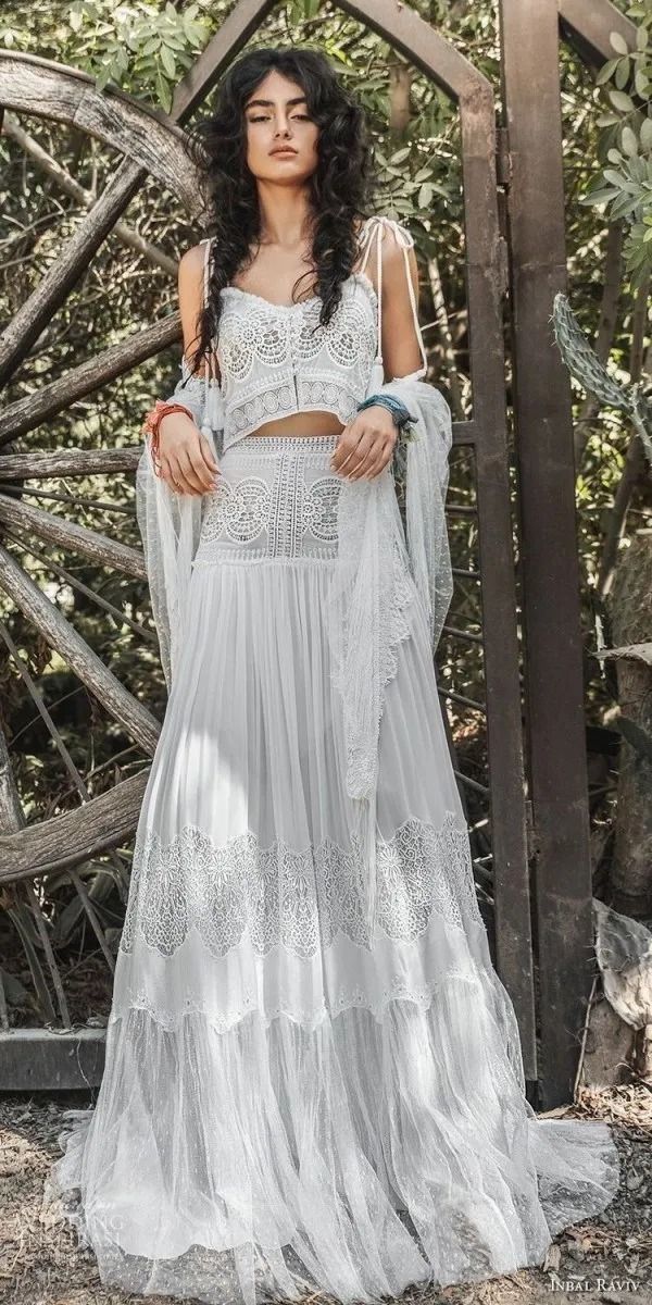 a woman wearing a white dress standing in front of a wooden structure with trees and bushes behind her