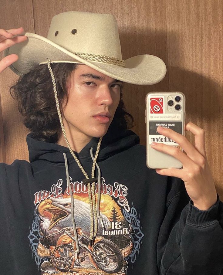 a young man wearing a cowboy hat and holding up his cell phone to take a selfie