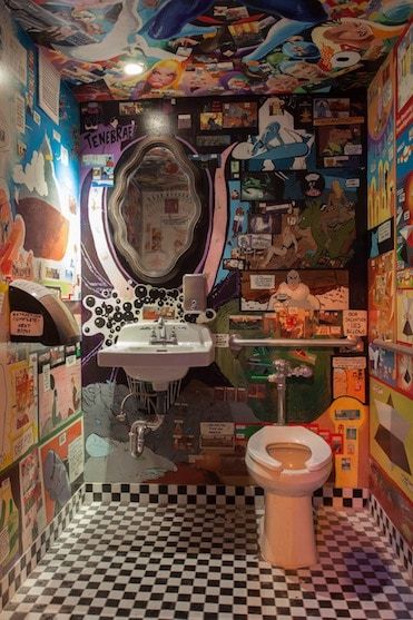 a bathroom with a toilet, sink and mirror covered in comic themed wallpapers