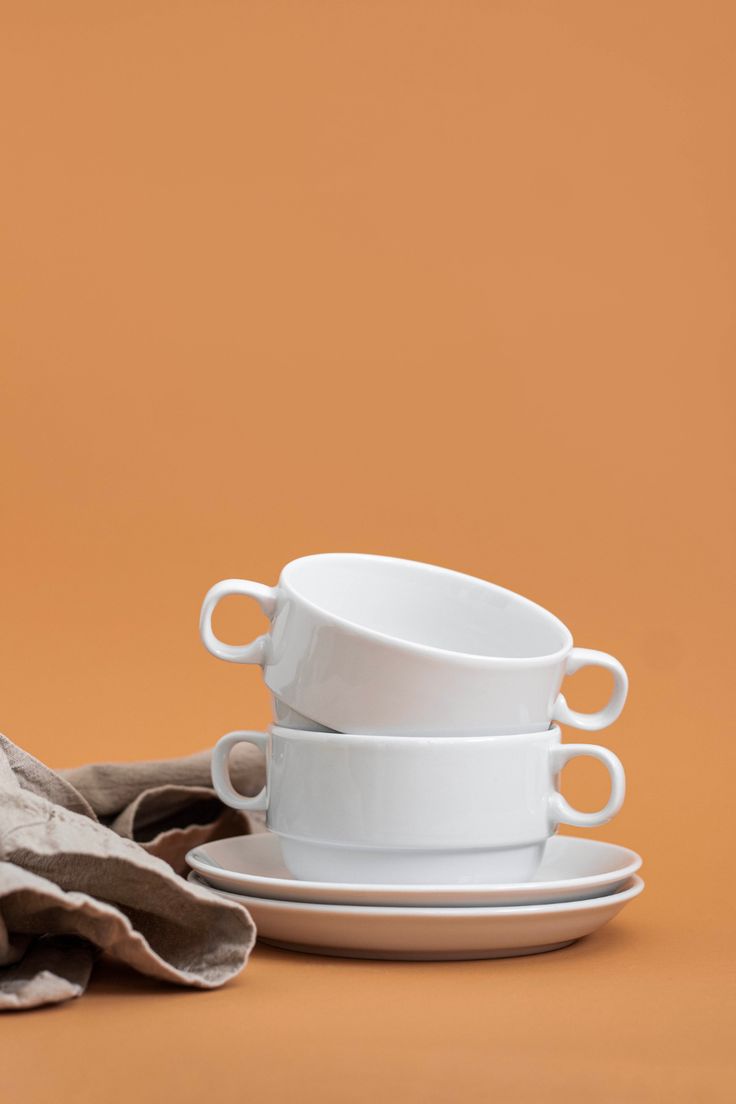 stack of white cups and saucers sitting on top of each other next to a brown cloth