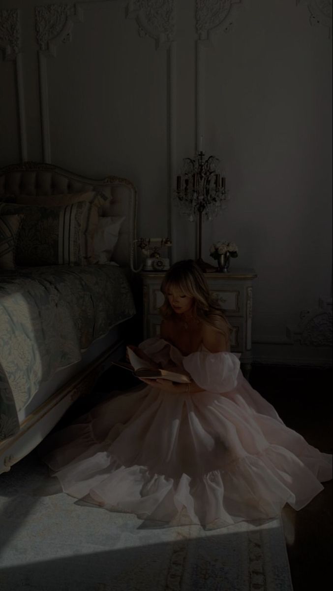 a woman in a dress sitting on the floor next to a bed and reading a book