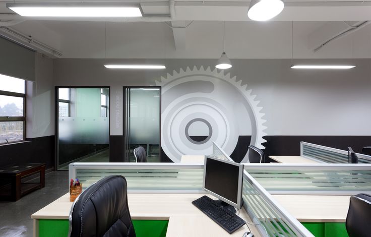 an office cubicle with desks, computers and large gear wheel on the wall