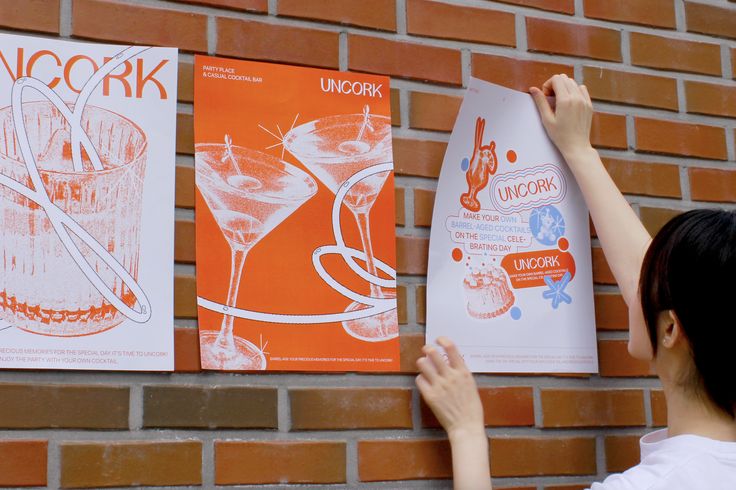 a woman holding up two posters on a brick wall next to a red brick wall