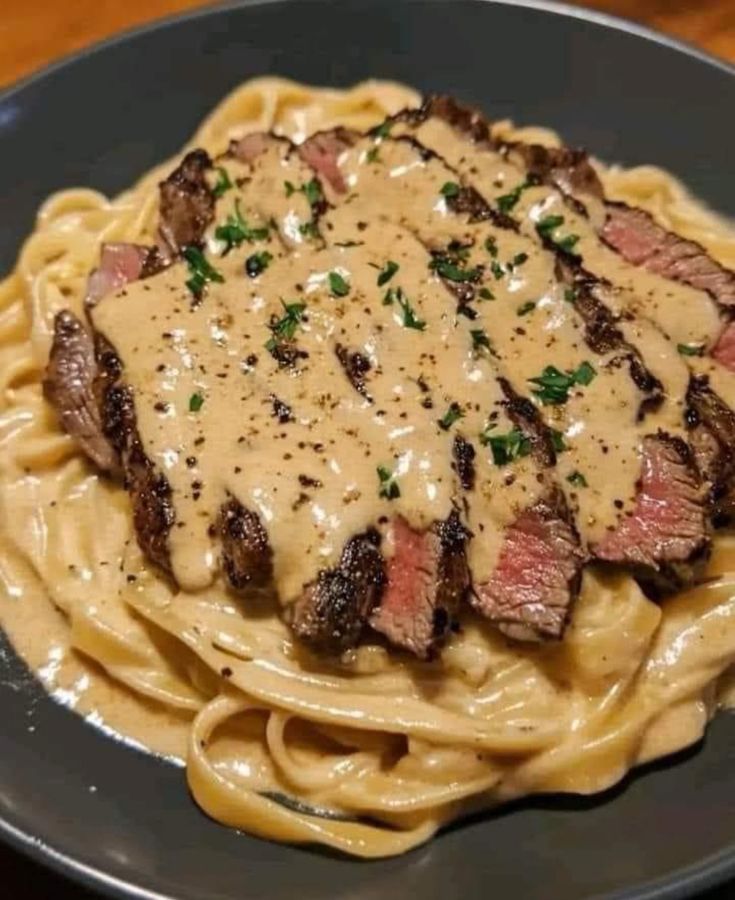 steak and sauce on top of pasta in a black plate with a wooden table behind it