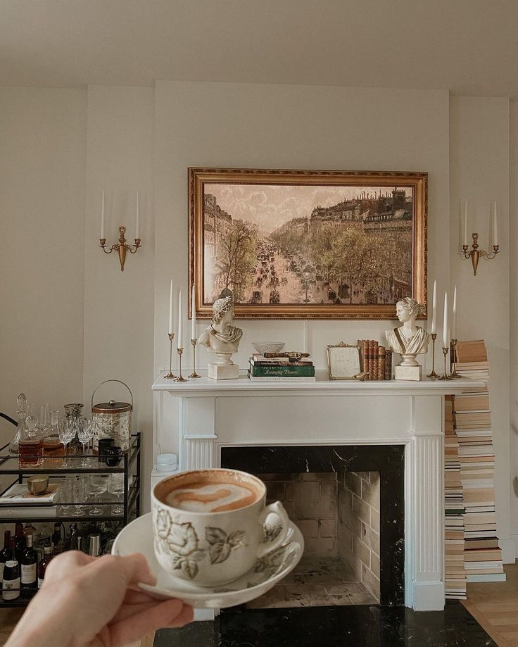 a person holding a coffee cup in front of a fire place with a painting on the wall