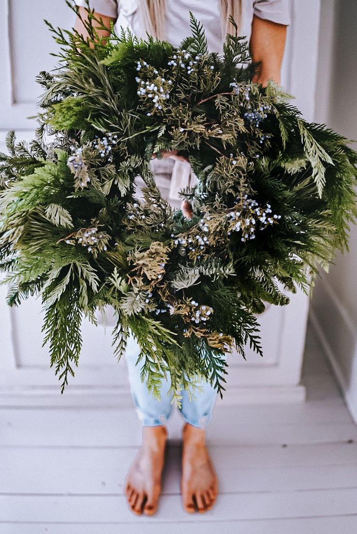 a person standing in front of a door holding a wreath with greenery on it