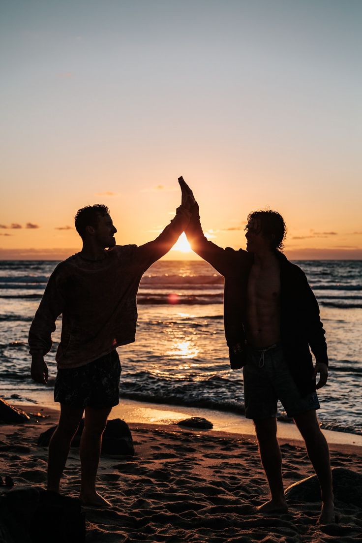 two men standing on top of a sandy beach holding their hands up in the air