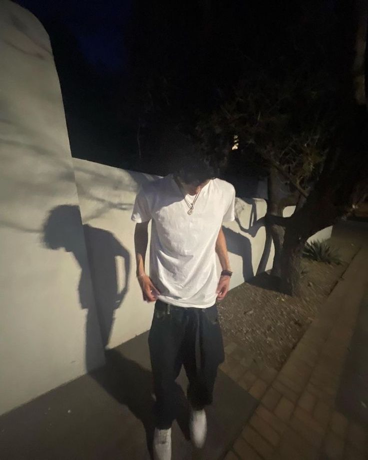 a man in white shirt and black pants standing on sidewalk next to tree at night