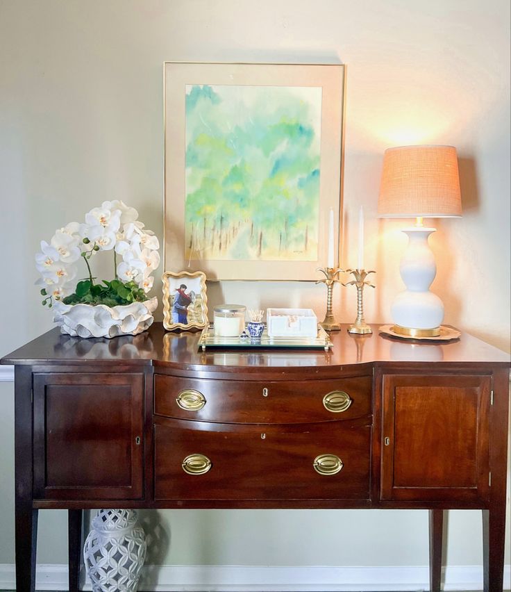 a wooden table topped with two lamps next to a painting and vase filled with flowers