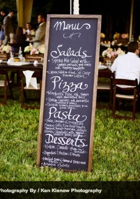 a chalkboard menu sign sitting on top of a grass covered field next to tables