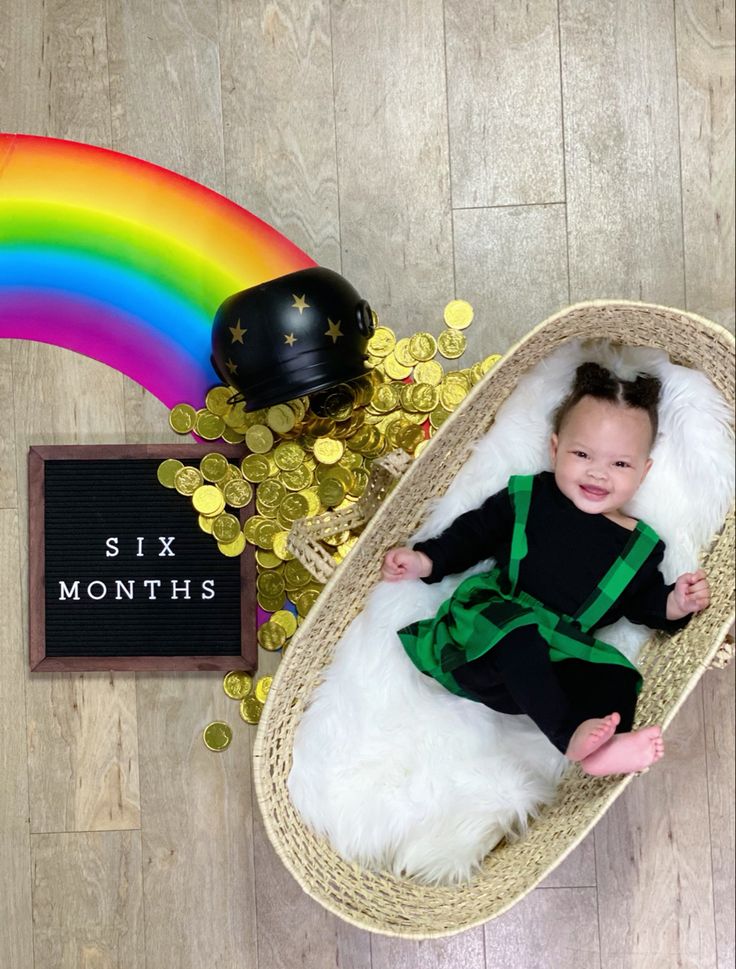 a baby sitting in a basket next to a rainbow and some coins on the floor