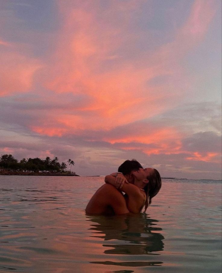 two people are kissing in the water with pink and blue clouds behind them at sunset