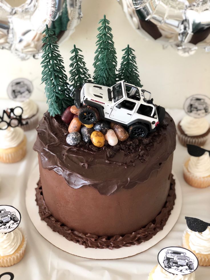 a chocolate frosted cake with cars and trees on top, surrounded by cupcakes