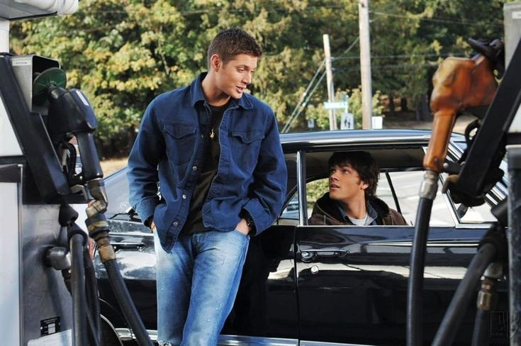 a man standing next to a car at a gas station with another man in the background