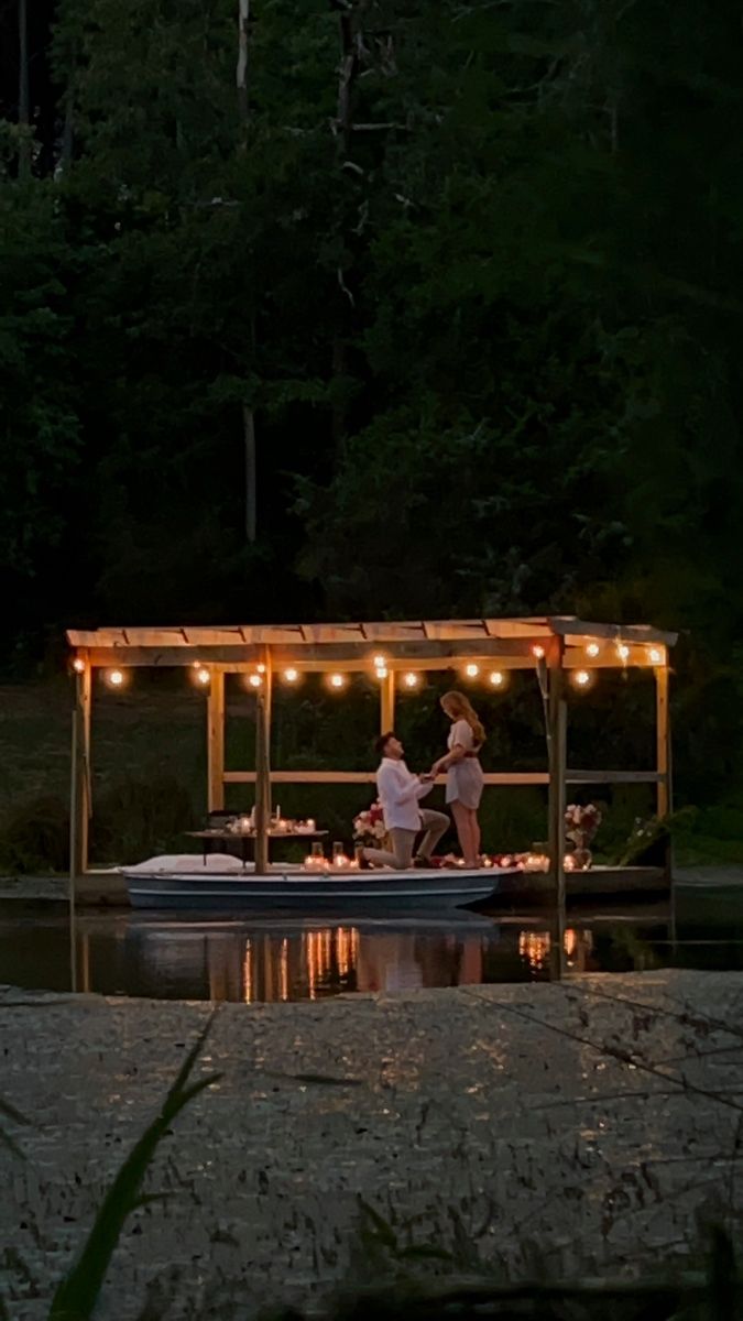 two people standing on a boat in the water at night with lights strung over it