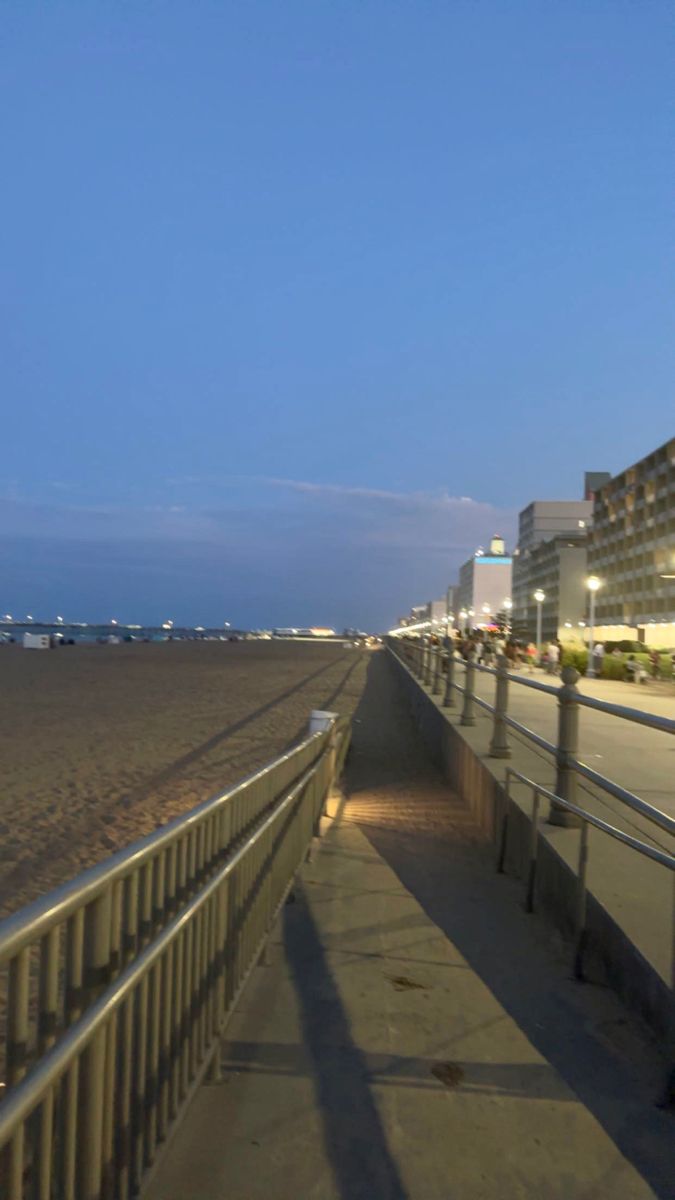 an empty sidewalk next to the beach at night with buildings in the background and lights on