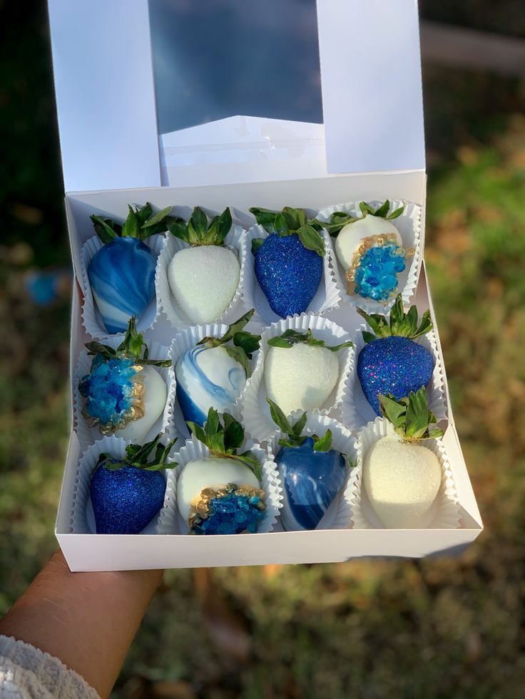 a hand holding an open box of chocolates with strawberries on them and blue decorations