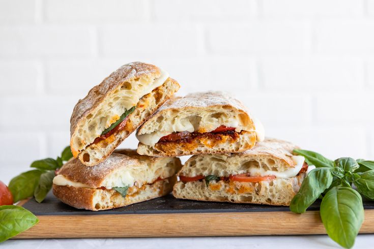 three sandwiches stacked on top of each other with fresh basil leaves around them and tomatoes in the background