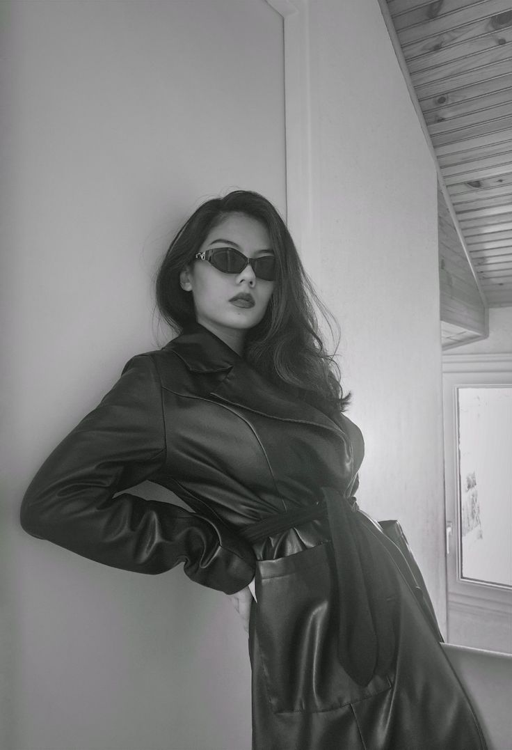 black and white photo of woman in leather coat leaning against wall with sunglasses on her face