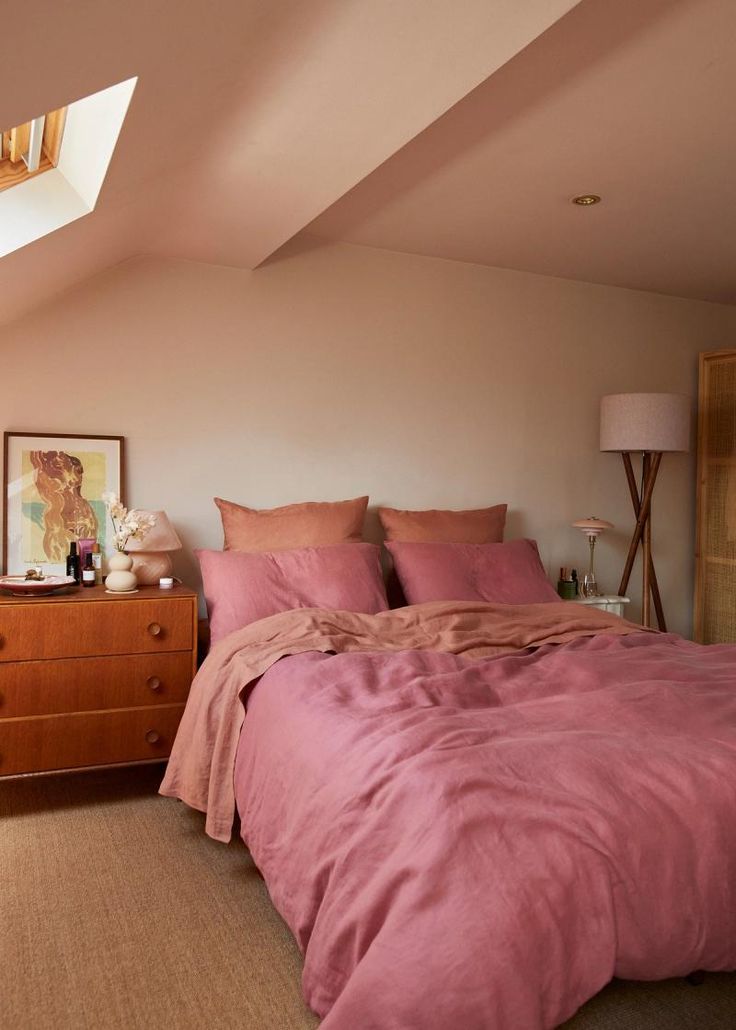 a bed with pink sheets and pillows in a bedroom next to a dresser, lamp and window