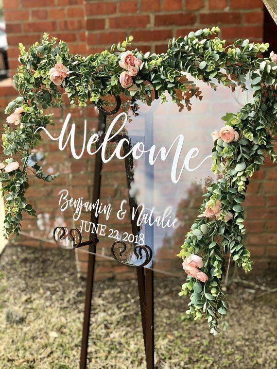 a welcome sign with pink flowers and greenery on the front, in front of a brick wall