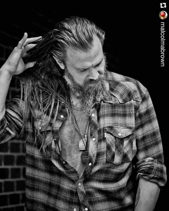 a man with long hair and beard wearing a flannel shirt is standing in front of a brick wall