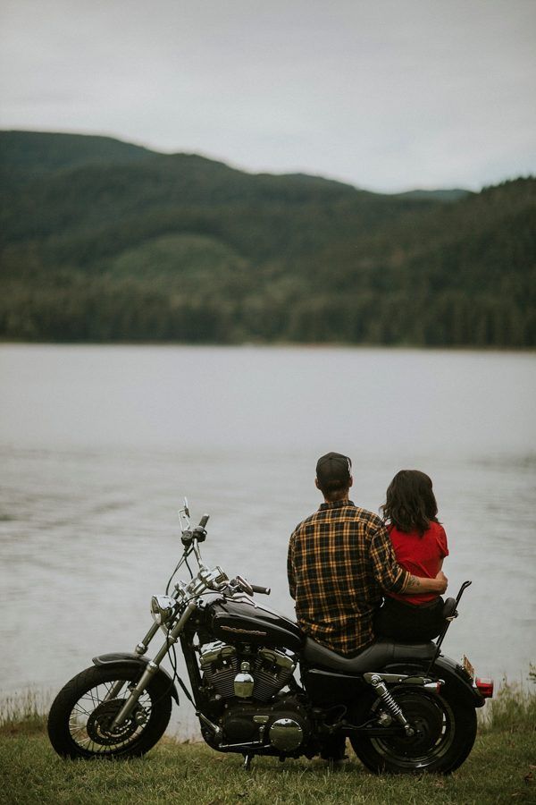 a man and woman sitting on a motorcycle looking out over the water with mountains in the background