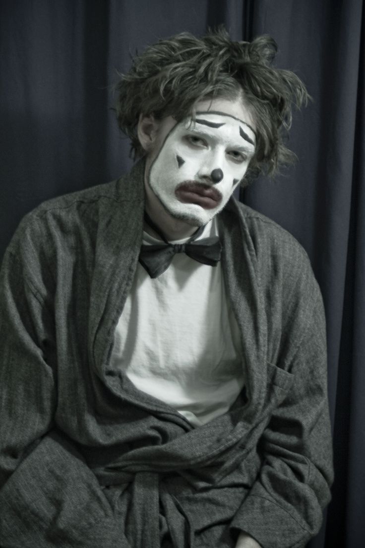 a man with clown makeup sitting in front of a black curtain wearing a gray robe