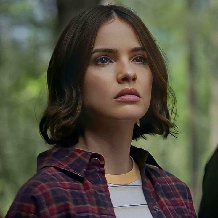 a woman with short hair wearing a plaid shirt and looking off into the distance while standing in front of trees