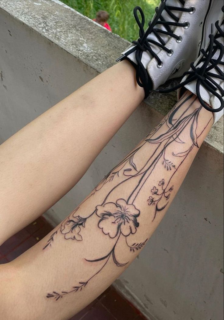 a woman's legs with flowers and leaves tattoo on her left leg, next to a wall