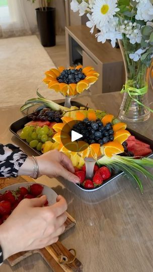 a woman cutting up fruit on top of a wooden table with flowers in the background