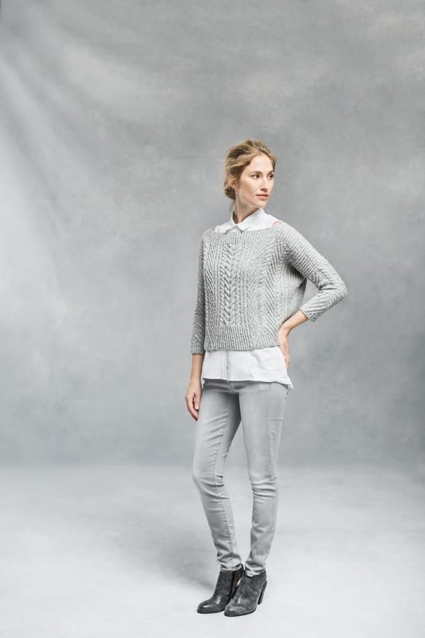 a woman standing in front of a gray background wearing grey pants and a white sweater