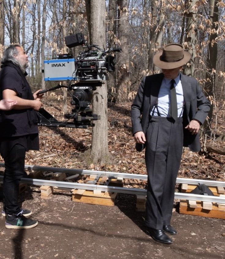 two men in suits and hats standing near a camera on a tripod with trees behind them