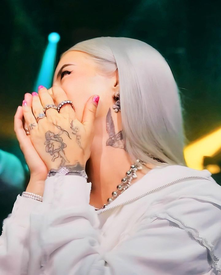 a woman with white hair and tattoos holding her hands up to her face while standing in front of a microphone